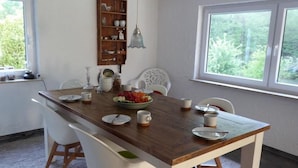 A dining area with a wooden table set for four, surrounded by white chairs. A wooden bookshelf with decorative items is mounted on the wall, and a window provides a view of greenery outside. - Vacation home time out from everyday life (Kirchenkirnberg)