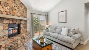 A living room with a stone fireplace, a grey sofa, and a view through a glass door to the outdoors.