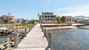 A wooden boardwalk leading to a waterfront property with a large house and a smaller building. - Dewey Sunset (Dewey Beach)