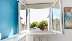 Una habitación con ventanal panorámico, radiador blanco y vista a un jardín con seto bien cuidado y un árbol.