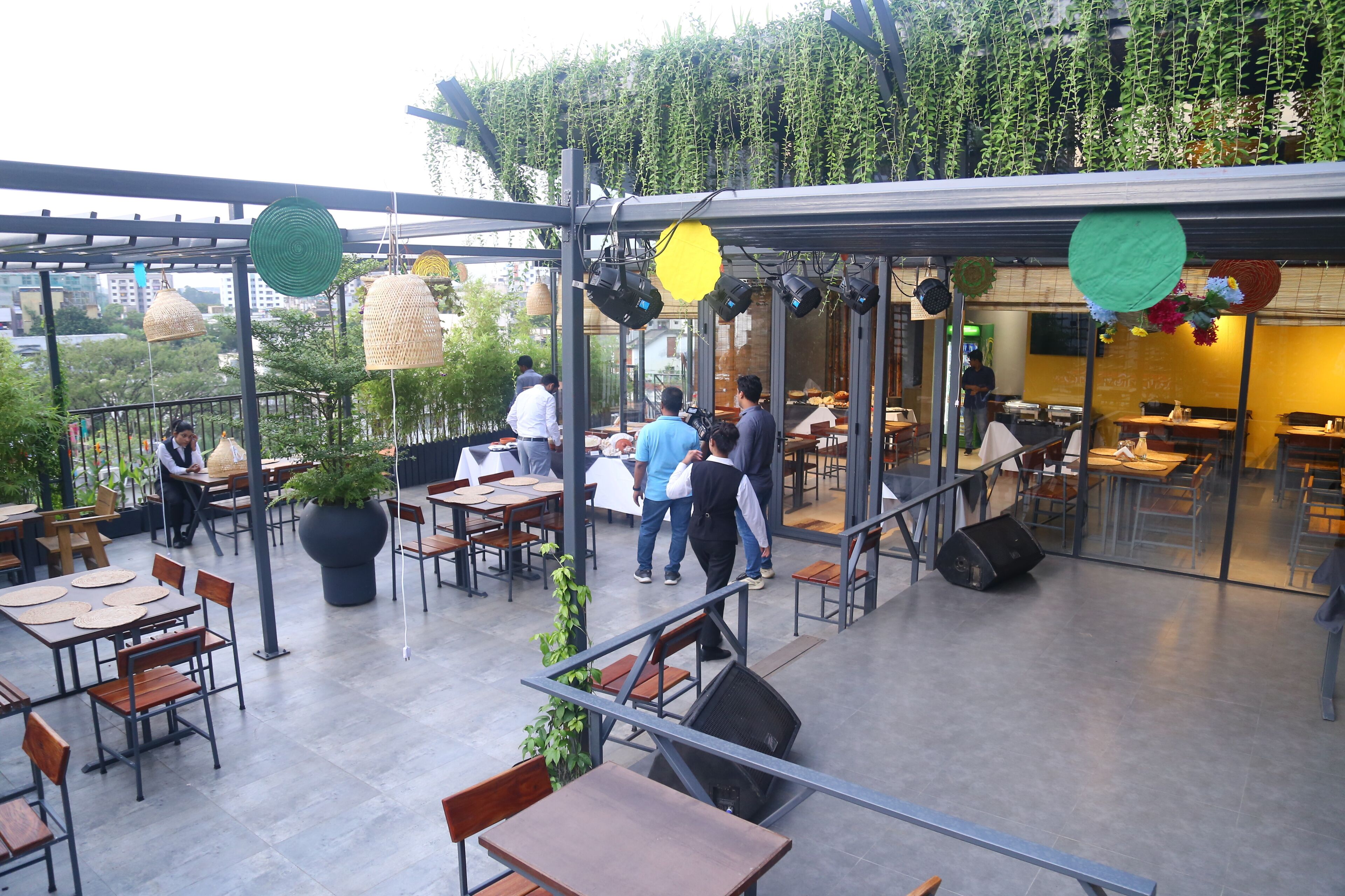 An outdoor dining area with tables and chairs, surrounded by greenery and a glass wall.