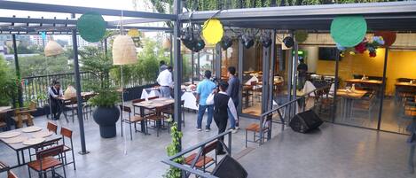 An outdoor dining area with tables and chairs, surrounded by greenery and a glass wall.