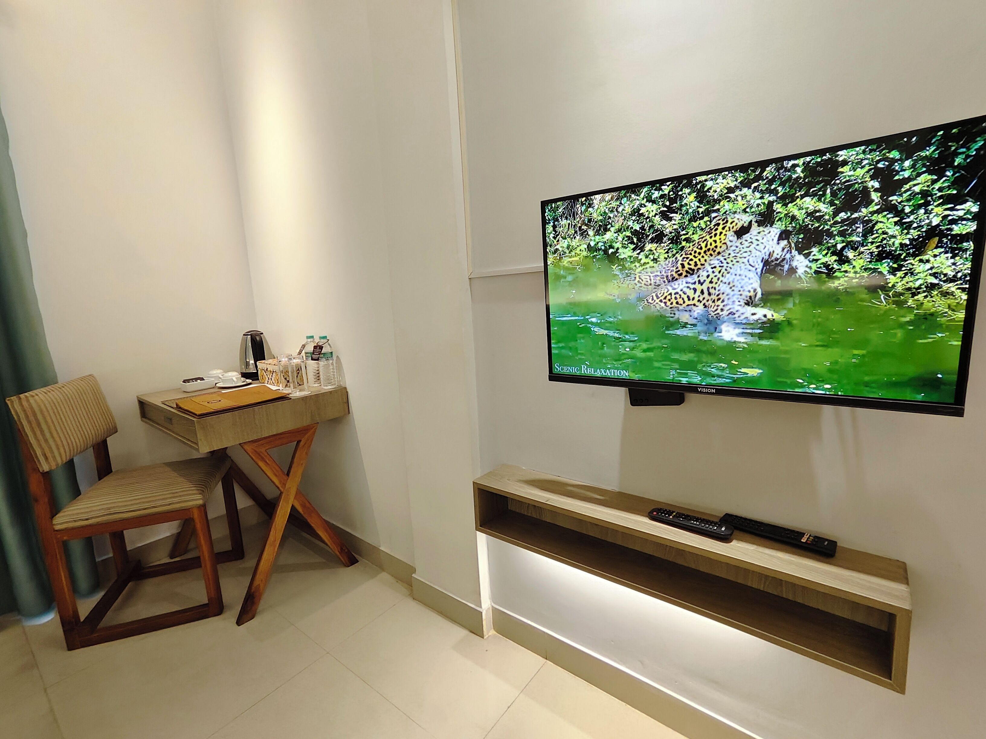A room with a TV mounted on the wall displaying a nature scene, a desk with a chair, and a shelf below the TV.