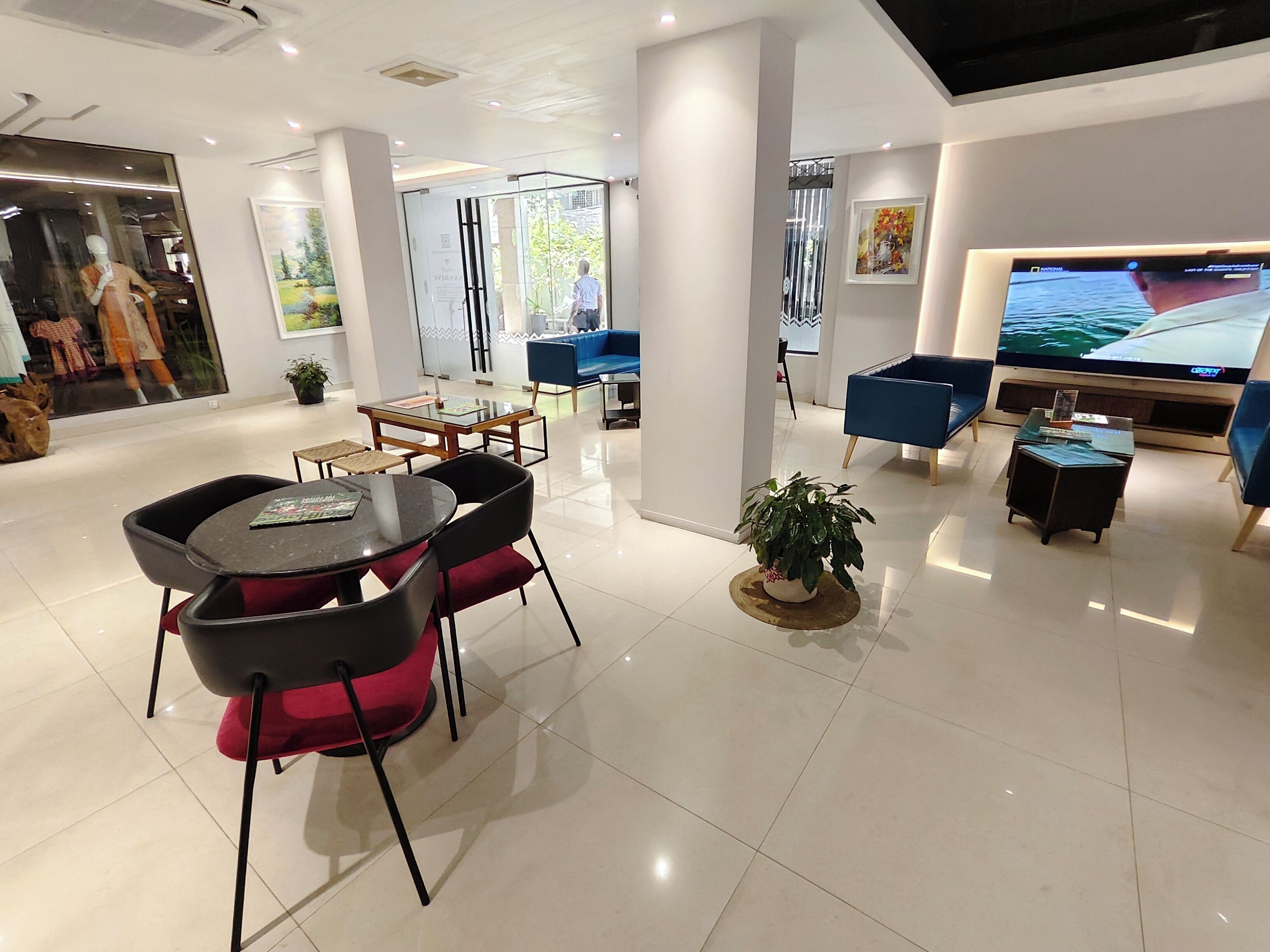 A modern hotel lobby with a seating area, a large TV screen, and a glass door leading outside.