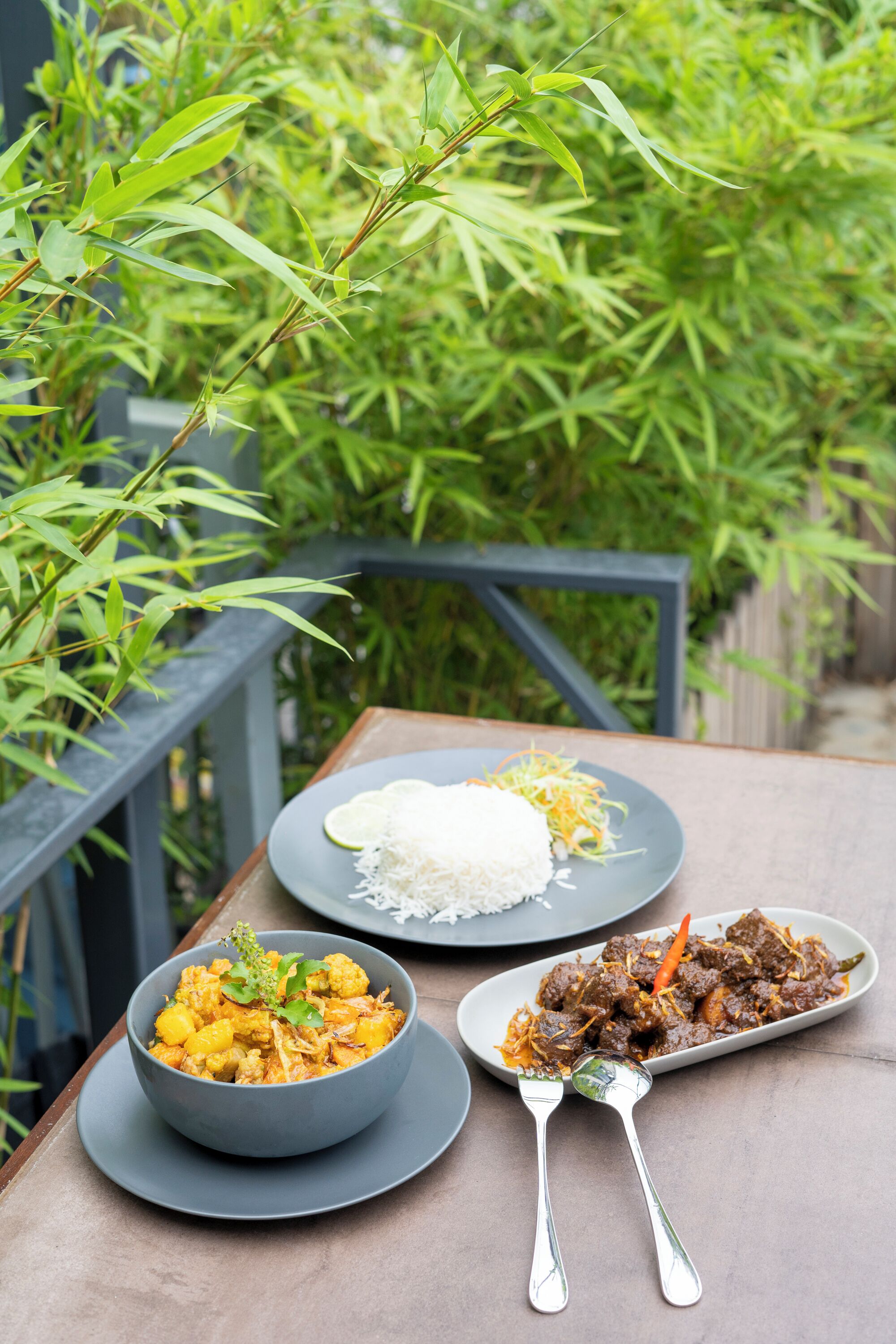 A table with two plates of food, one with white rice and another with a meat dish, set outdoors with bamboo plants in the background.