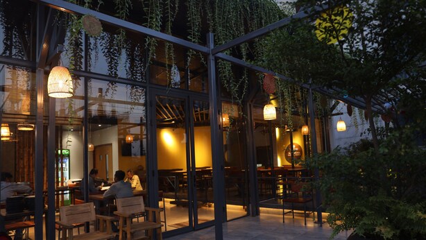 An indoor dining area with wooden tables and chairs, glass partitions, hanging plants, and pendant lighting. - Hotel Nandini (Dhaka)