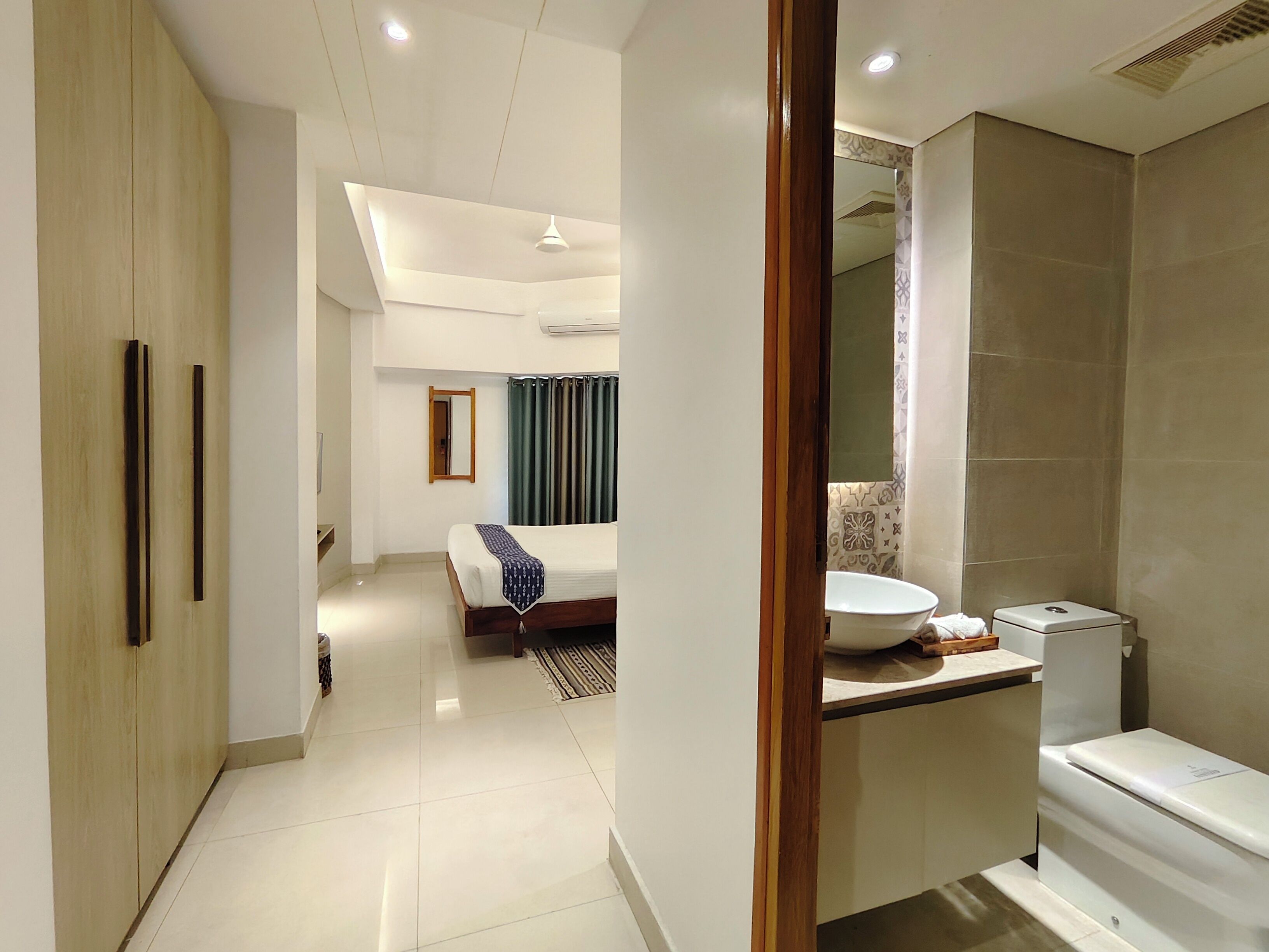A modern hotel room with a large bed, a bathroom with a sink and toilet, and a hallway leading to another room.
