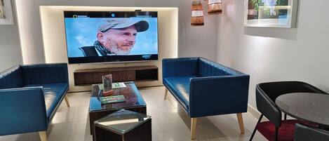 Una sala de estar moderna con televisor de pantalla plana, sofás tapizados en azul, una mesa de centro de vidrio e iluminación colgante.