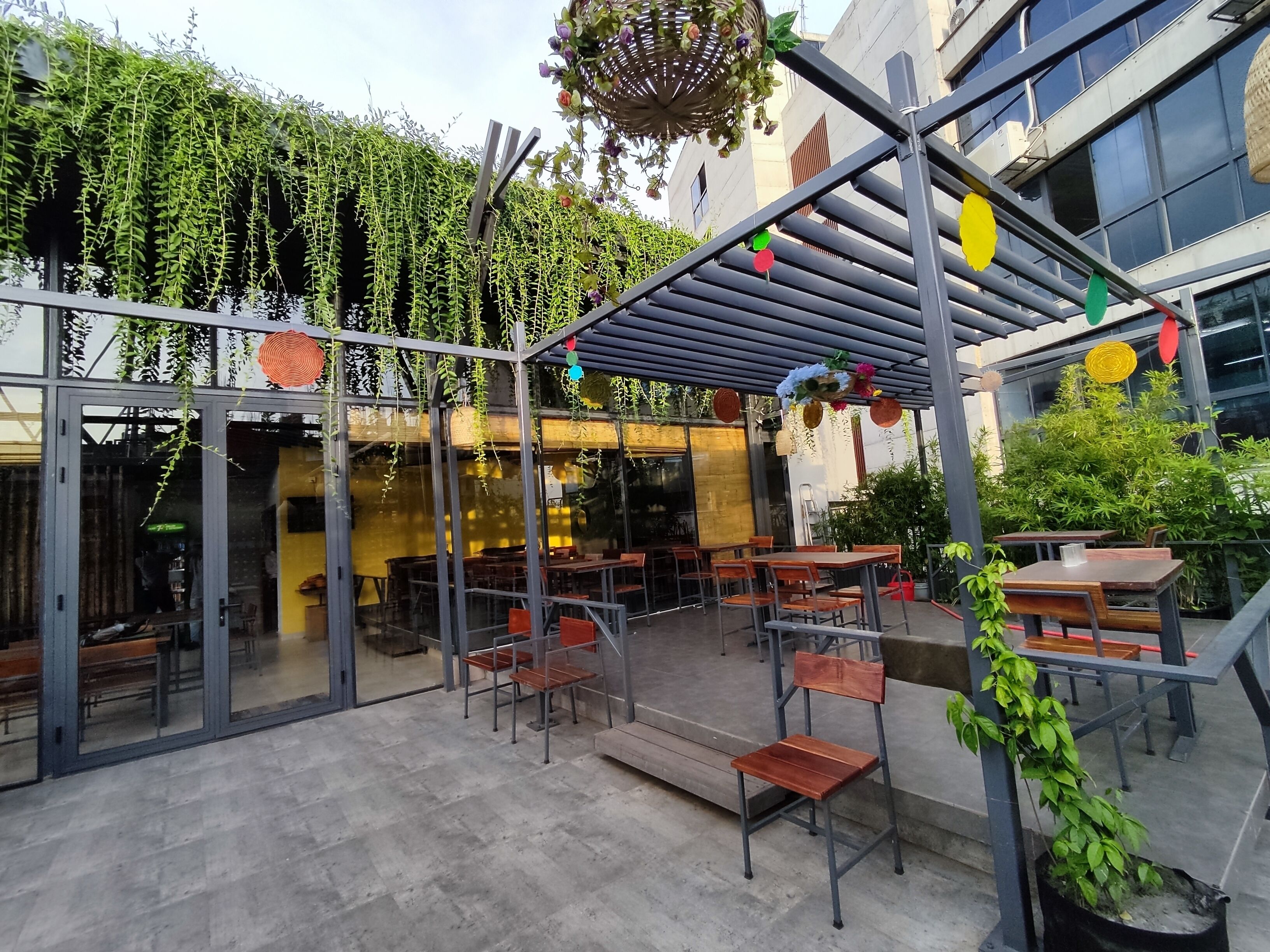 Ein Außenbereich mit Terrasse, Tischen und Stühlen, einer Pergola mit hängenden Dekorationen und Blick auf ein Gebäude mit großen Fenstern.