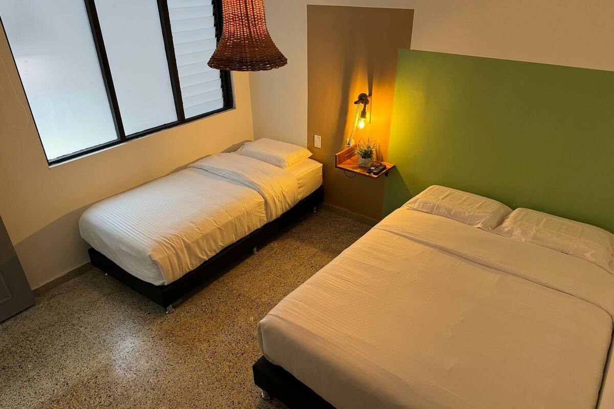 Une chambre avec deux lits, une fenêtre avec des stores, un luminaire suspendu et une étagère fixée au mur, sur laquelle se trouve une plante.