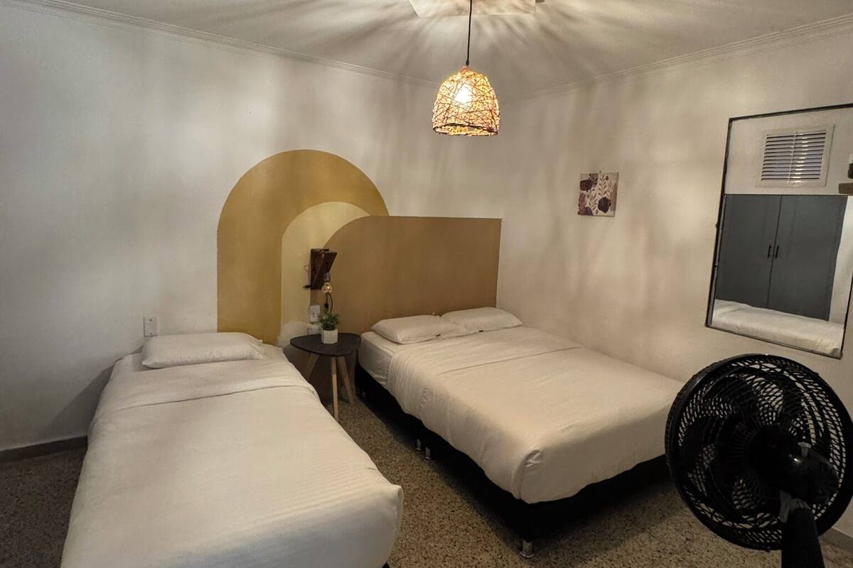 Une petite pièce bien éclairée, avec deux lits simples, une table, un ventilateur et un luminaire au plafond original.