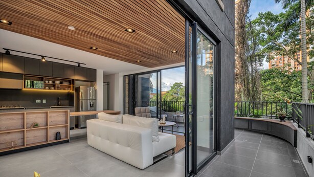Un salon moderne avec un canapé blanc, un coin cuisine doté d’étagères intégrées et une grande porte vitrée donnant sur un balcon extérieur.