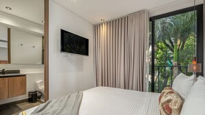 Une chambre d’hôtel moderne dotée d’un grand lit, d’un téléviseur fixé au mur, d’une salle de bain avec des toilettes et un lavabo, et offrant une vue sur les arbres par la fenêtre.