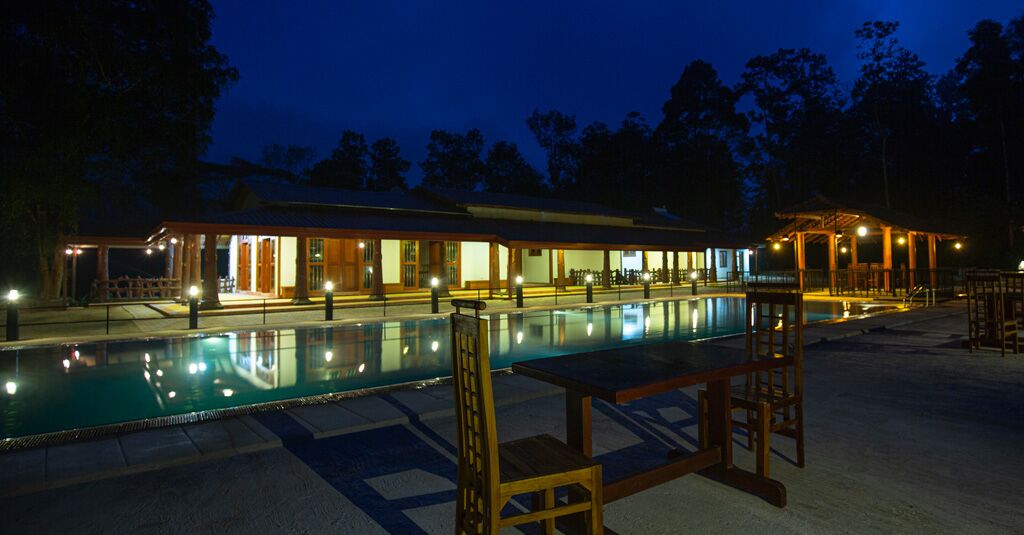 Vista nocturna de un hotel con piscina, mobiliario exterior y una zona cubierta con iluminación.