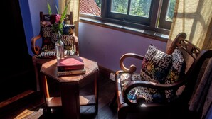 A cozy room with a wooden chair and a small table, a window with a view of trees, and a patterned cushion on the chair.