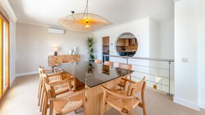 A modern dining area with a glass table, wooden chairs, a unique woven light fixture, and a kitchen visible through an archway. - Villa Maya Luxurious House With Pool by LovelyStay (Praia da Luz)