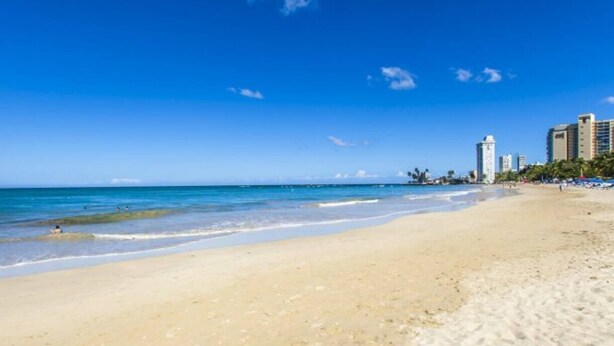 A beach with clear blue water, sandy shore, and high-rise buildings in the background. - GIVB-Updated Family Budget unit just step away from the beach! (Carolina)