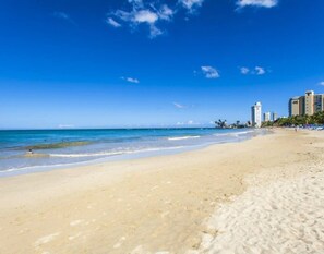 Ein Strand mit klarem, blauem Wasser, sandigem Ufer und Hochhäusern im Hintergrund.
