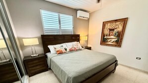A bedroom with a bed, nightstands, a window with blinds, an air conditioner, and a wall-mounted painting. - GIVB-Updated Family Budget unit just step away from the beach! (Carolina)
