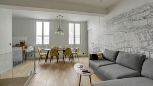 A modern living room with a grey sofa, a dining area with yellow chairs, and a large wall mural of a cityscape. - 168 Suite M - Superb apartment in Paris (Paris 02 Bourse)