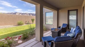 Eine überdachte Terrasse mit Korbmöbeln, einem Feuerkorb und Blick auf einen angelegten Garten und Berge.