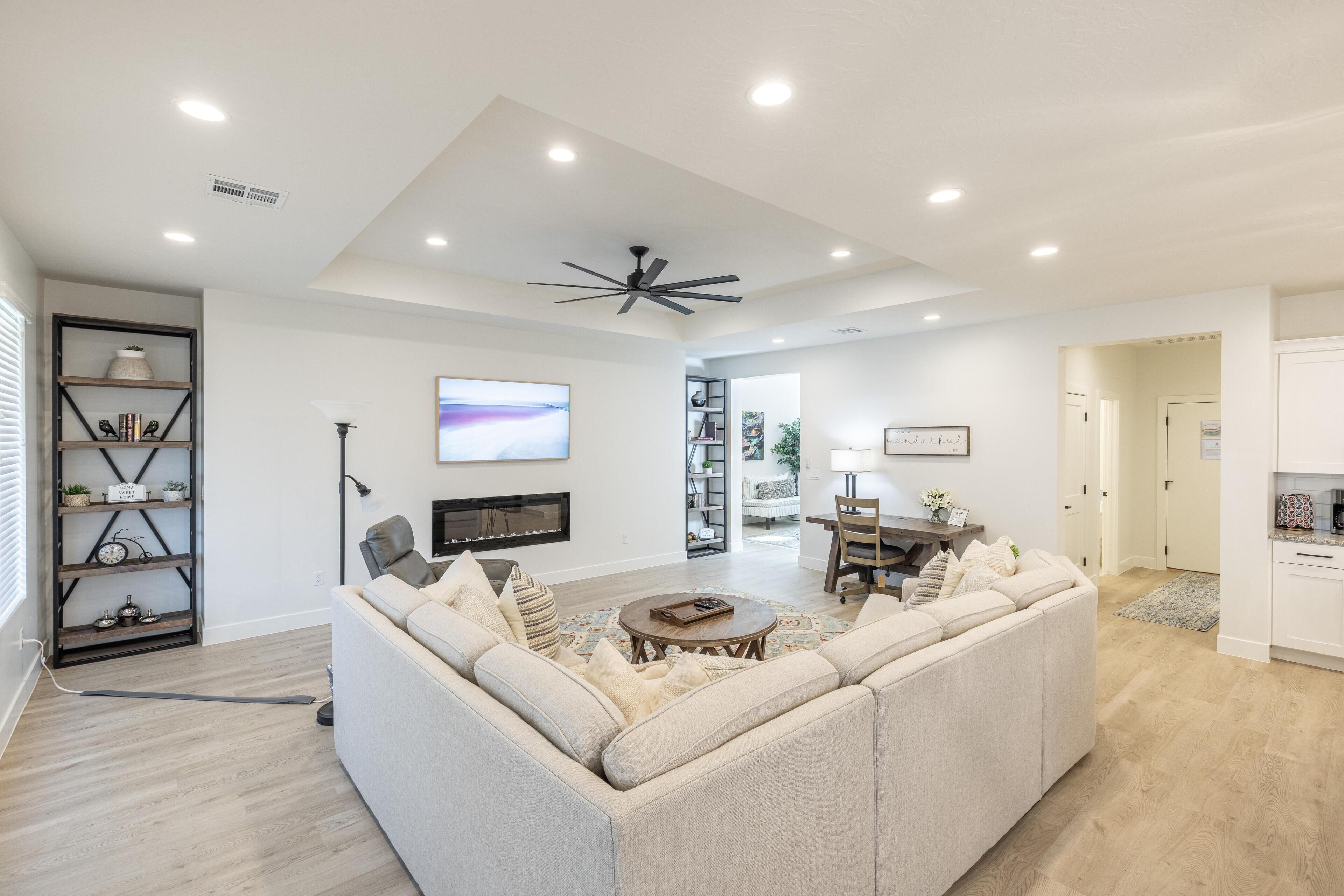 A modern living room with a sectional sofa, a coffee table, and a ceiling fan. There is a TV mounted on the wall and a bookshelf with decorative items.