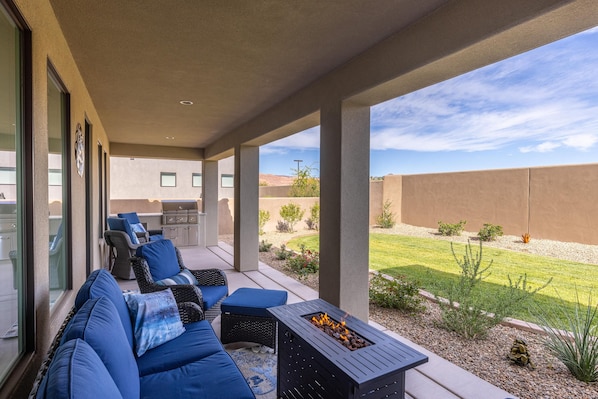 A covered patio with outdoor seating, a fire feature, and a view of a landscaped yard.