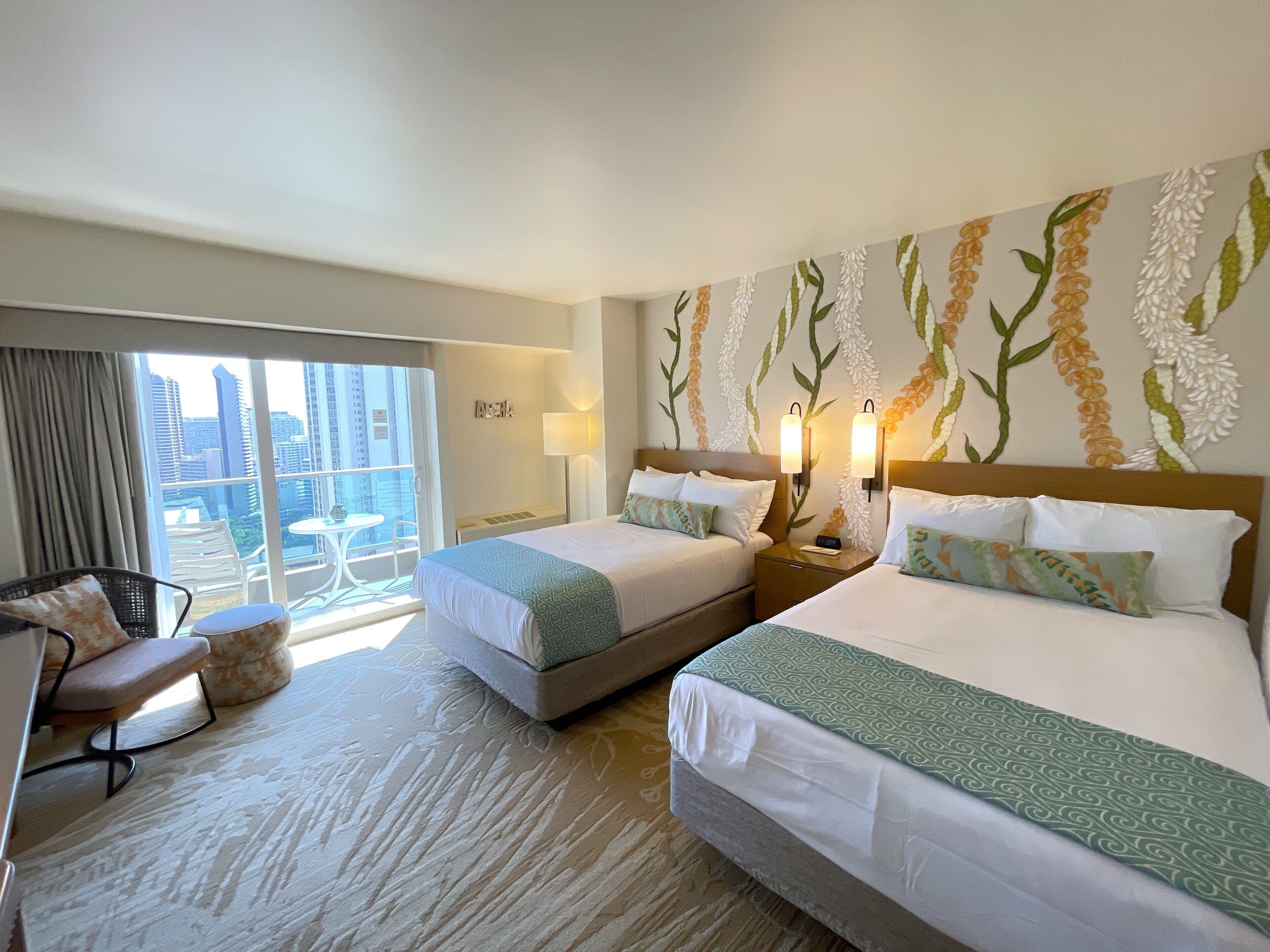A hotel room with two beds, a balcony with a view of the city, and a small table with chairs.