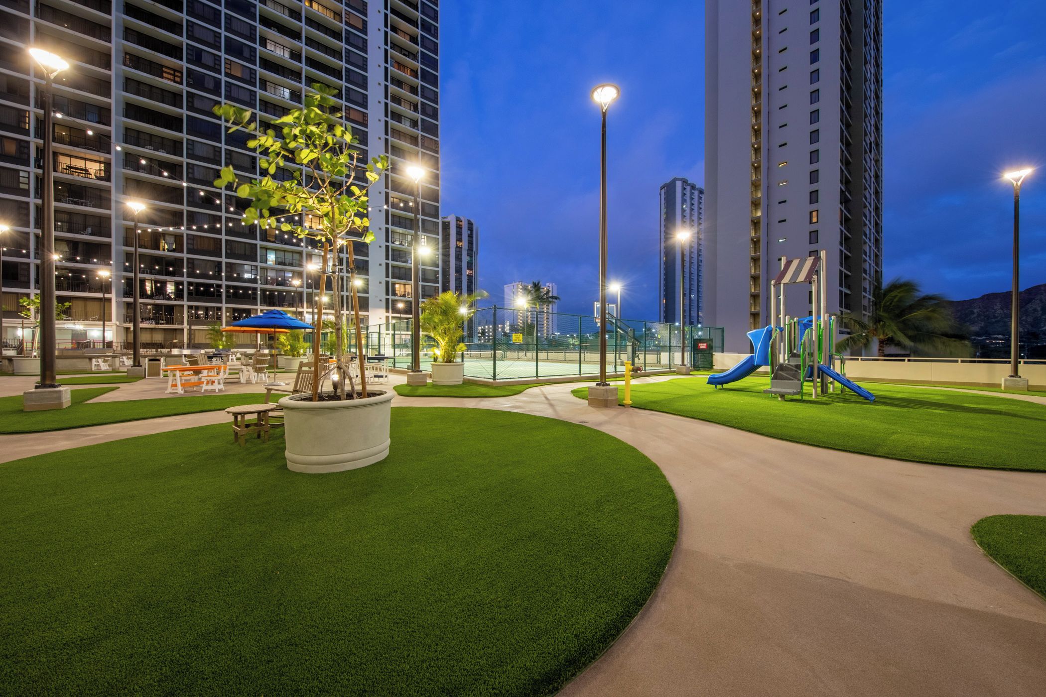 A landscaped outdoor area with a playground, benches, and a large building in the background.