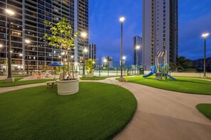 A landscaped outdoor area with a playground, benches, and a large building in the background.