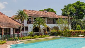 Un área de piscina con una piscina azul cristalina, un edificio con techo de tejas y un jardín cuidado con palmeras y arbustos.