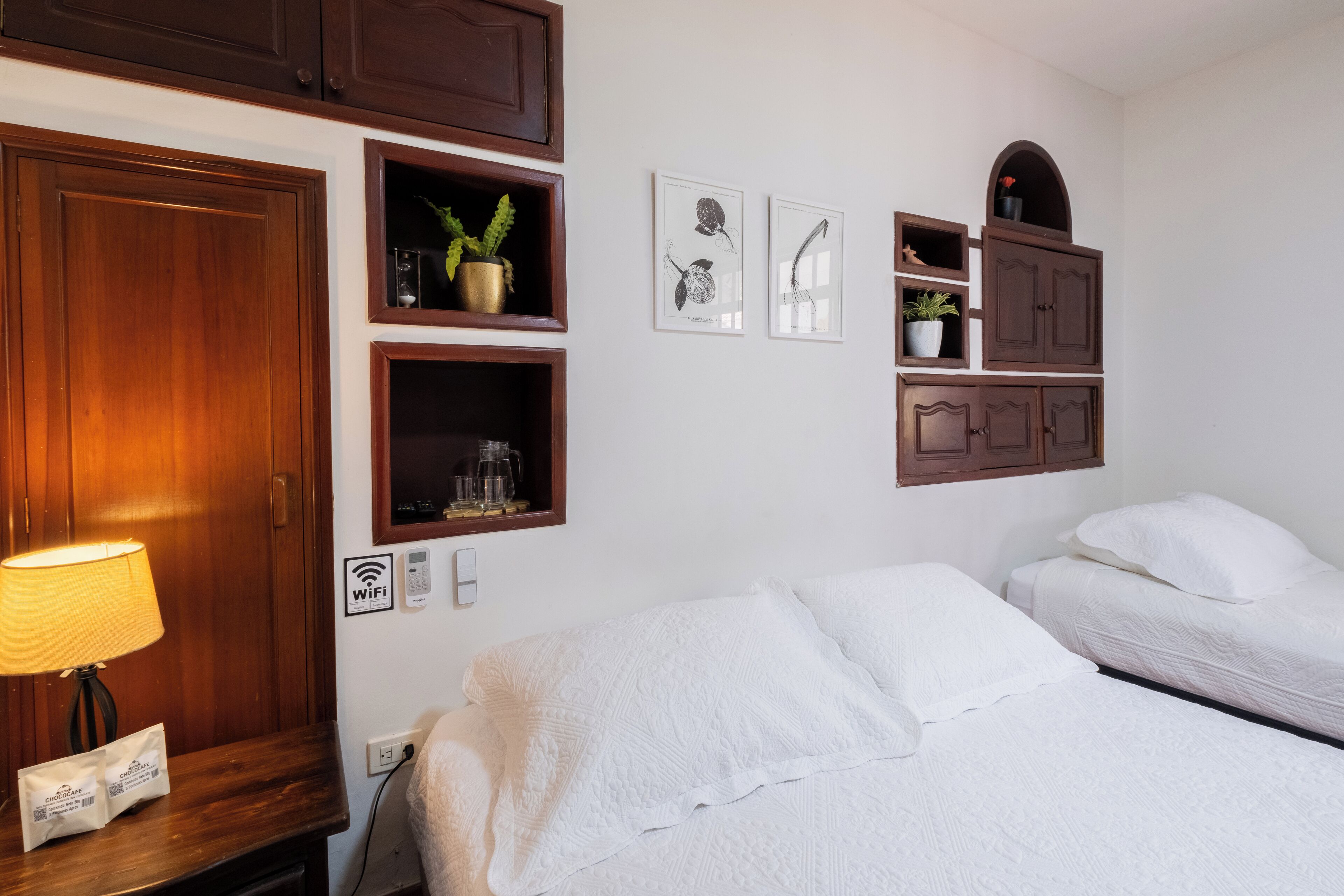 Quarto de hotel com um guarda-roupa de madeira, mesa de cabeceira com abajur, cama branca e estante com plantas e quadros.