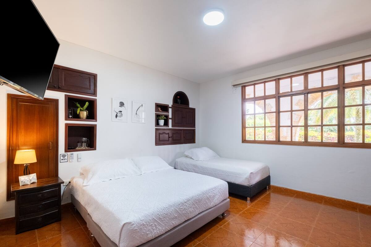 Una habitació d'hotel amb dos llits, televisió, escriptori amb làmpada i finestra amb vista a vegetació.