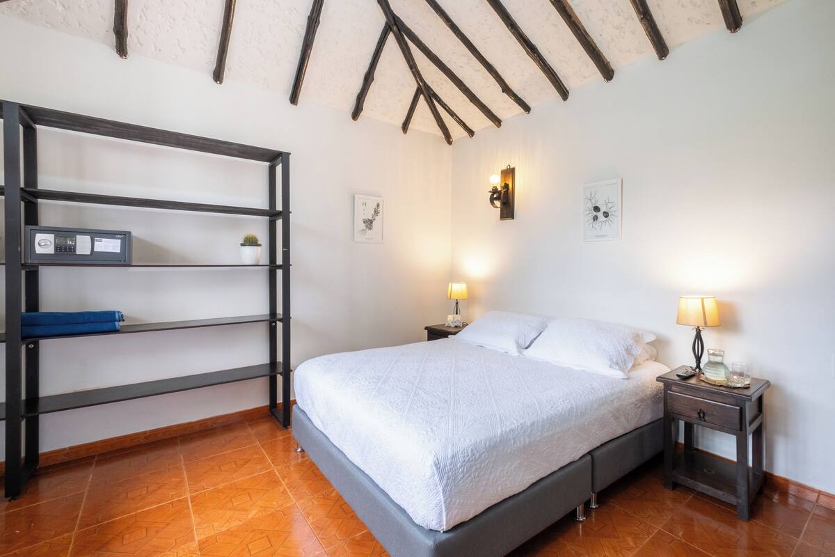Un dormitori amb un llit, una tauleta de nit, un prestatge i un sostre amb bigues a la vista.