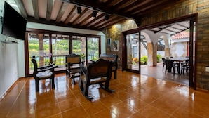 Un patio amplio con mobiliario de madera, un televisor y vistas al jardín a través de puertas de vidrio.