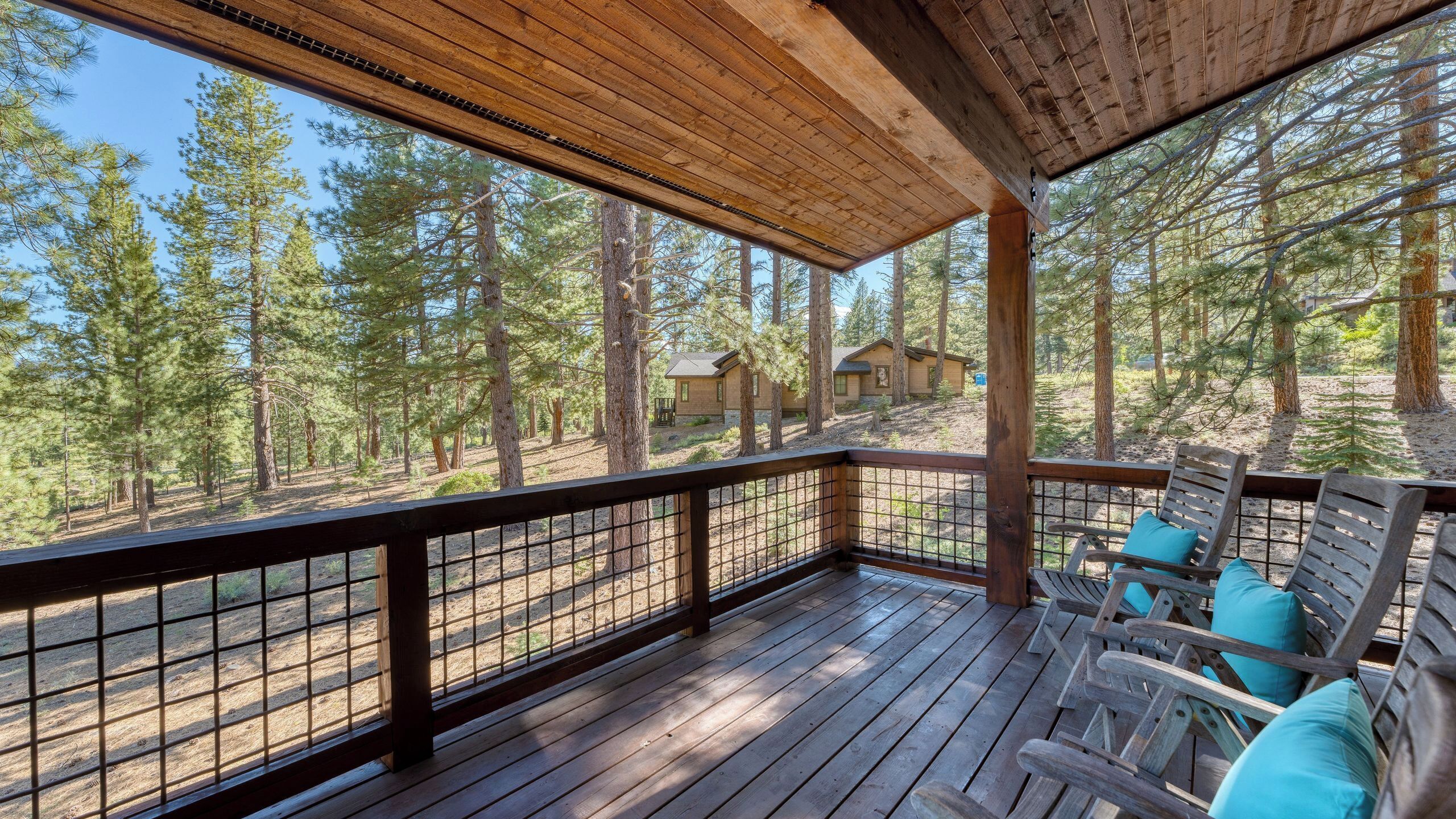 A wooden deck with a railing, wooden chairs with cushions, and a view of a forest with cabins.