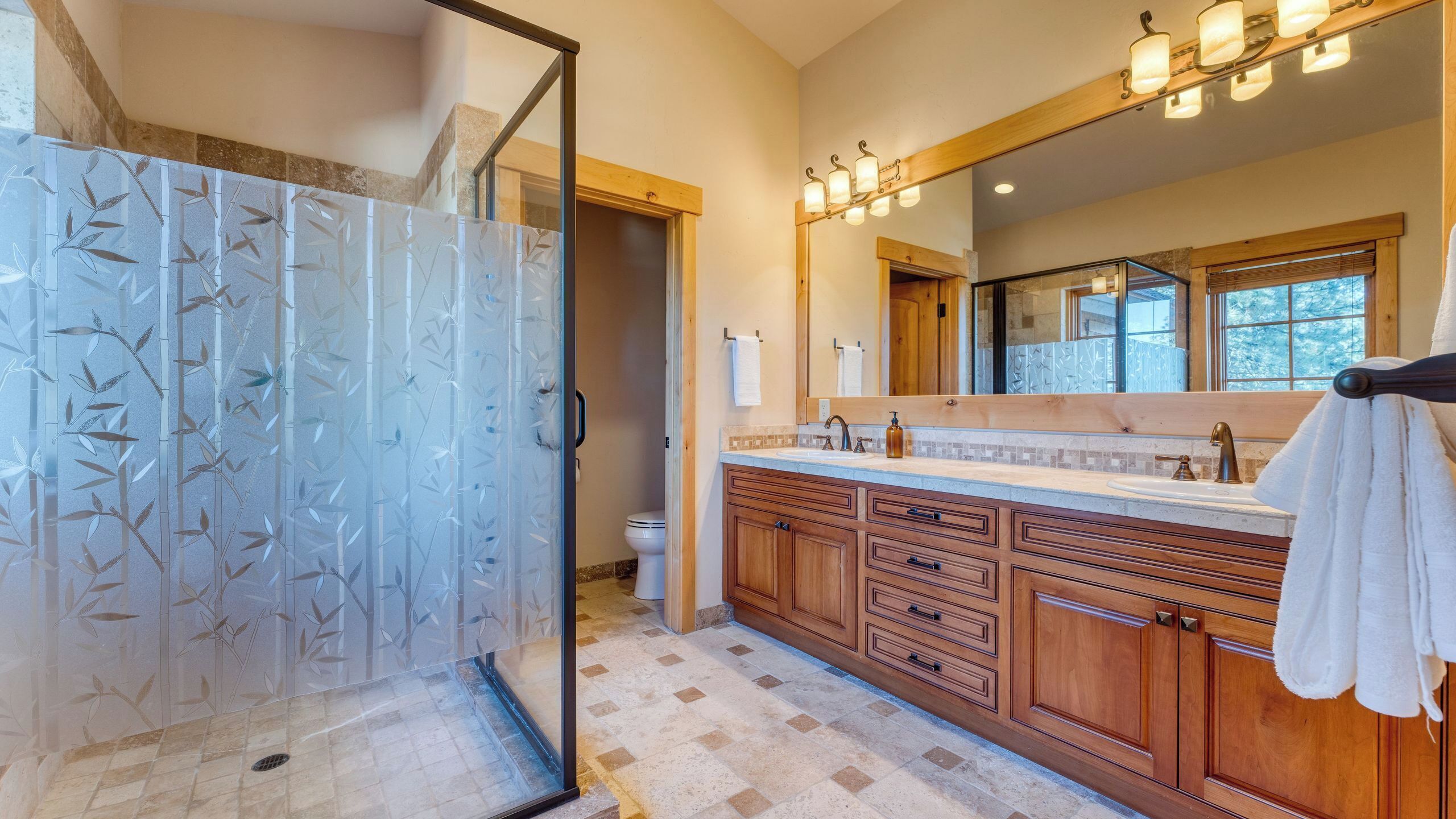 A bathroom with a glass shower enclosure, wooden cabinets, a large mirror, and a window.