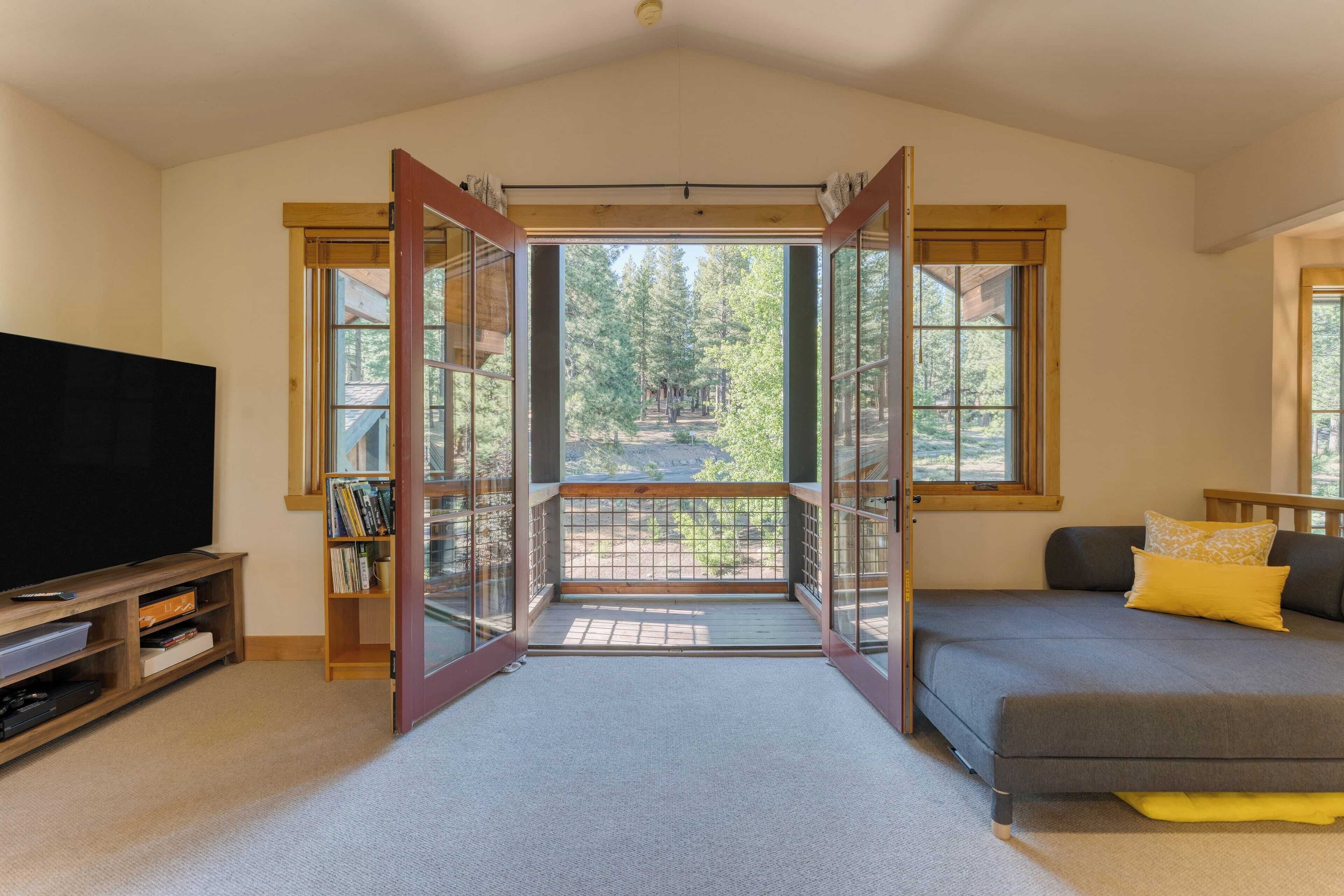 A room with a large TV, a bookshelf, a grey sofa, and a balcony with a view of trees.