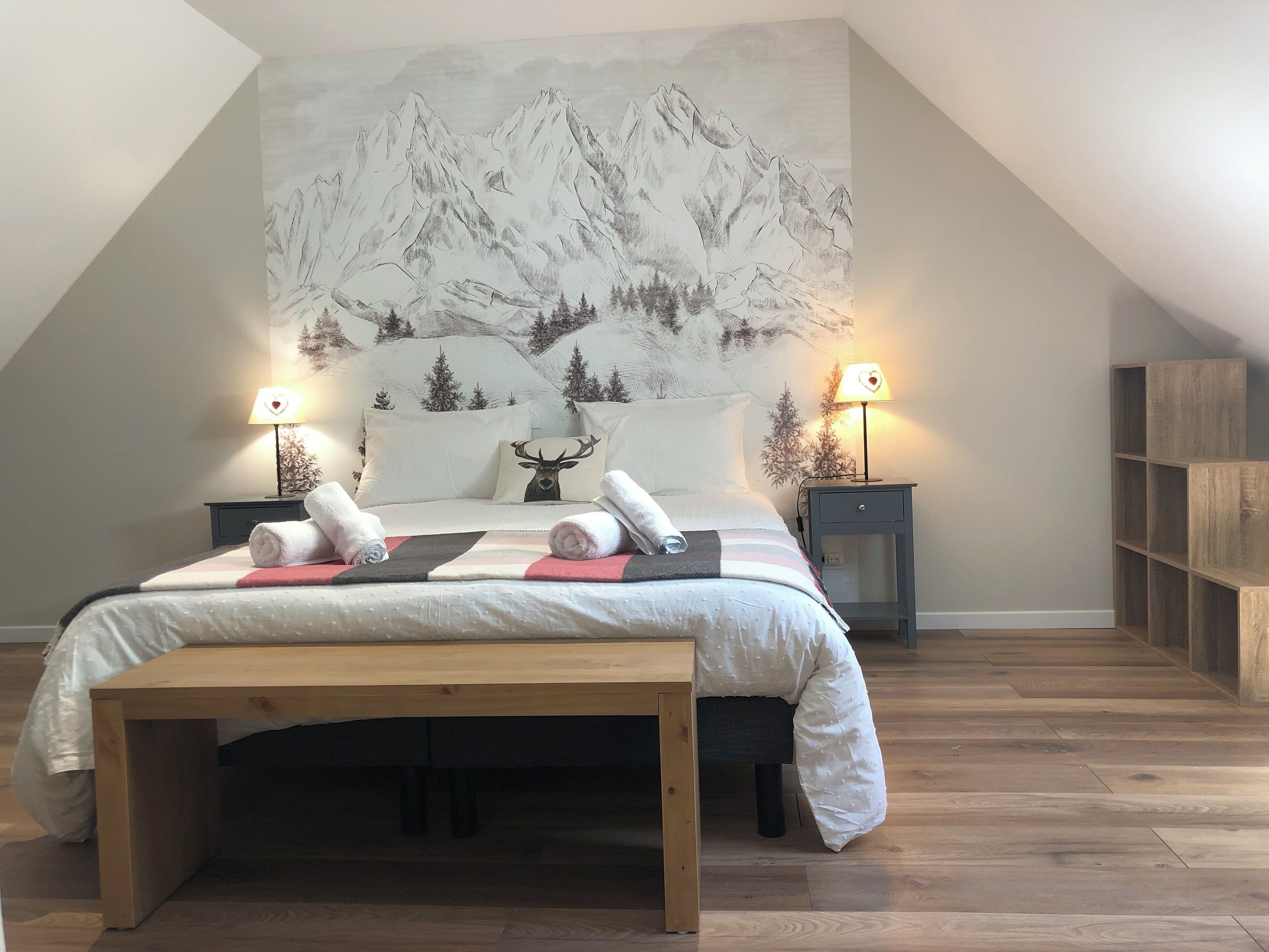 A bedroom with a bed, two bedside tables, a wooden bench, a wall-mounted lamp, and a bookshelf.