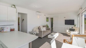 Ein modernes Wohnzimmer mit Sofa, Couchtisch und Fernseher. Es gibt eine Küchenzeile mit weißen Schränken und einem Esstisch.