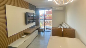 Una sala moderna con televisor de pantalla plana, paneles de madera, un comedor con mesa y sillas, y un ventanal con vistas a edificios.