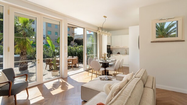 A spacious living area with a sofa, dining table, and chairs, large sliding glass doors leading to a balcony with plants and a view of buildings. - Casa Amare 4+2, Emma Villas (Camaiore)
