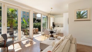 A spacious living area with a sofa, dining table, and chairs, large sliding glass doors leading to a balcony with plants and a view of buildings. - Casa Amare 4+2, Emma Villas (Camaiore)