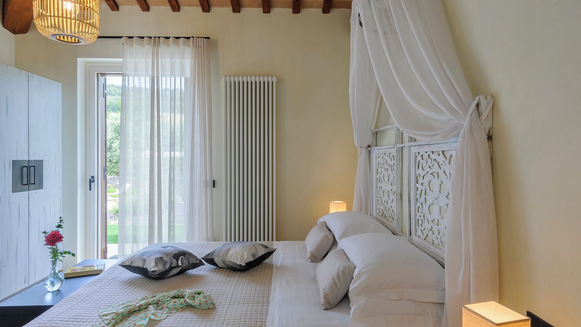 Ein Schlafzimmer mit Bett, Nachttisch, Lampe, einem Fenster mit Vorhängen und Blick ins Grüne.