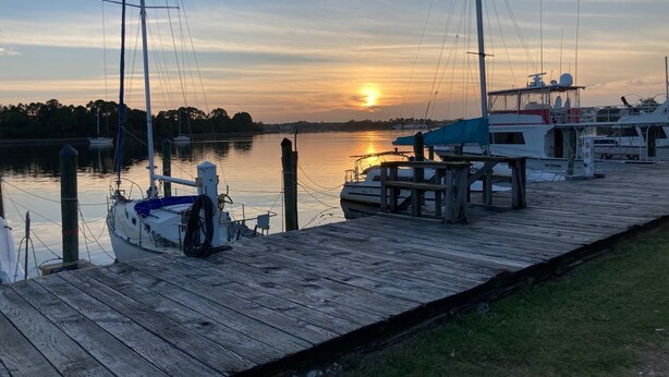 Un muelle de madera con barcos amarrados al final, un cielo despejado y una puesta de sol al fondo.