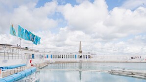 Une piscine avec un long bassin rectiligne et, en arrière-plan, un bâtiment surmonté d’un monument imposant.