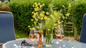 Une table ronde en verre, avec une bouteille de vin et des verres, entourée de chaises bleues, installée à l’extérieur, avec une haie en arrière-plan.