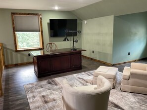 A spacious living room with a dark wooden desk, a television mounted on the wall, and a large window providing natural light. - Secluded and spacious Barndominium close to 2 Ski Resorts and many attractions! (Factoryville)