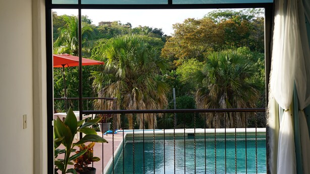 Vista desde un balcón con piscina, rodeado de exuberante vegetación y un paraguas rojo.
