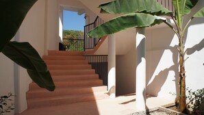 Una escalera conduce a un balcón con vista a árboles, una pared blanca y una planta grande.
